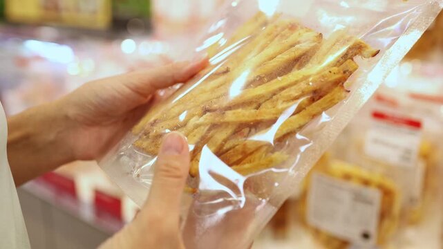 Close-up of a woman hands holding and choosing a vacuum-sealed plastic package of dried fish strips or snacks from display shelves in a grocery store or supermarket. Retail shopping, food.