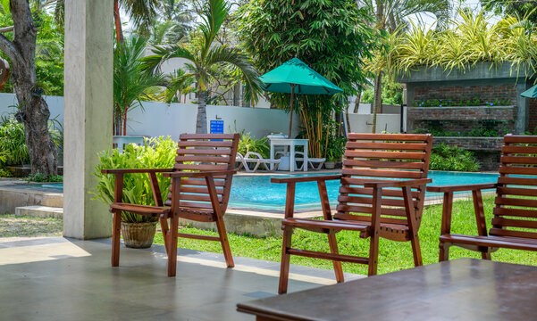 Wooden chairs overlook a clear resort swimming pool, tropical plants, sun loungers, umbrellas, and landscaped greenery