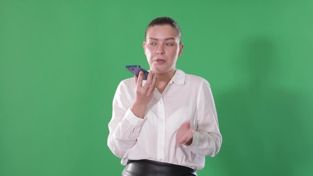 Young woman stands against the background of a chroma key with a mobile phone and records an audio message in a messenger. European woman communicates on a mobile phone on a green background. High