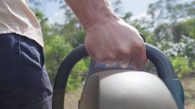 POV view from a camera mounted to a petrol chainsaw as carried toward Australian bushland. The chainsaw is visible in the foreground as the man approaches fallen timber and deadfall