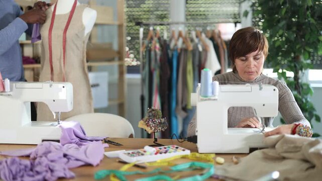 Woman tailor sews clothes in sewing machine. Worker uses sewing machine to create clothes for clients. African male companion work in background with mannequin 