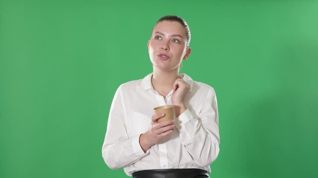 Young woman in a white blouse and a black skirt holds a cup of coffee to take away against the background of chrome key, she smiles happily and enjoys a delicious drink . High quality 4k footage