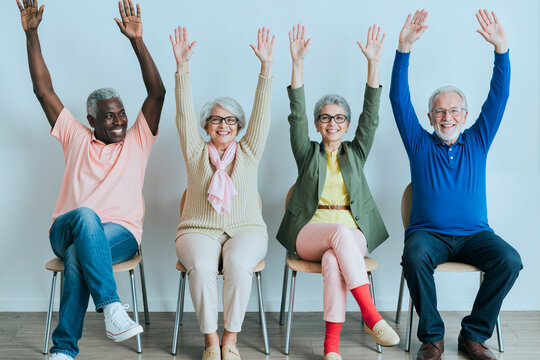 Wide shot of diverse seniors sitting in row and raising hands during group session, showing participation and active aging. Ideal for healthcare, retirement, community support marketing