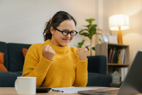 Asian businesswoman happily celebrates online success looking at laptop screen from home office. Excited female entrepreneur clenches her fists in celebration of victory and good news.