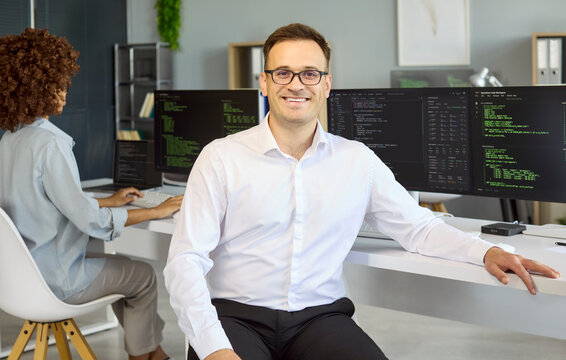 Male programmer developer coding software on computer. Smiling man with glasses sits by monitors in a clean office, code visible and coworker behind. Concept of efficient software teamwork.