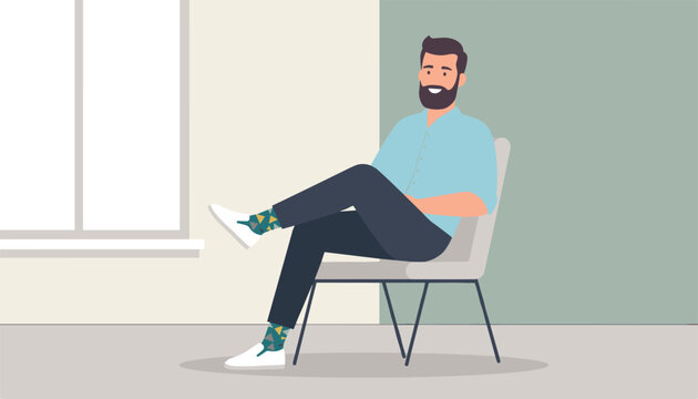 Bearded man in blue shirt and dark trousers sits relaxed on a modern chair with legs crossed at the ankle.
