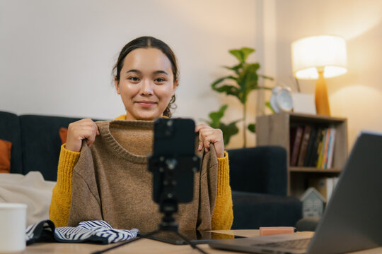 Asian female online seller live streaming clothes sale via smartphone at home. Happy woman showing sweater sweater fashion product to customer via mobile camera for social media e-commerce.