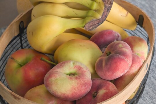 Fresh fruits in a wooden basket including apples, peaches, and bananas on a simple gray background