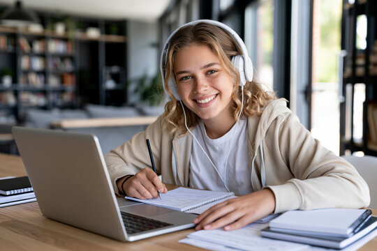 Young girl smiles while studying at a desk. She wears headphones and writes in a notebook. Laptop sits beside her, open and ready. Natural light fills the bright, modern room. Perfect for educational