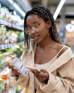 Young woman examines milk carton with smartphone. She wears round glasses and braided hair. Her expression shows curiosity and focus. Grocery aisle shelves display various products