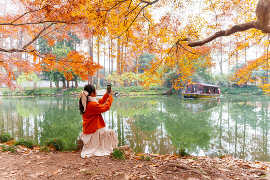 Female traveler holding smartphone taking pictures of a classic wooden boat on calm lake surrounded by autumn foliage in Hangzhou, China