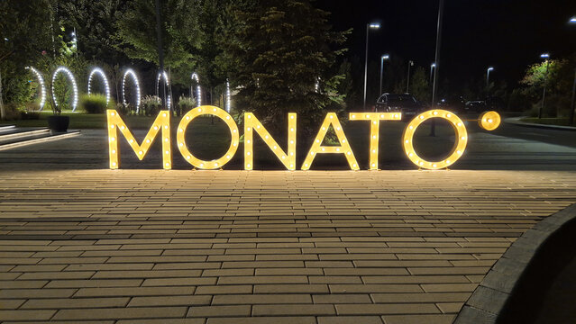 Illuminated glowing letters spell "MONATO" on a brick plaza at night, surrounded by trees and decorative lighting. Picture taken on August 23, 2026 in Ilfov, Romania.