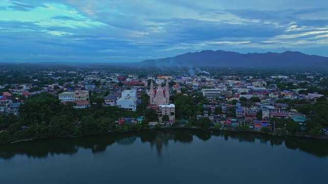 Wide establishing drone footage of Iglesia Ni Cristo near Sampaloc Lake in San Pablo City, Laguna, showcasing urban neighborhoods, tropical greenery, and scenic mountain views during twilight.