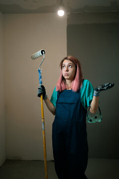 
Puzzled Female Painter Looking Away Holding White Roller Brush. Thoughtful young woman decorator wearing blue overalls holds painting roller looks sideways curiously
