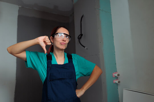 Dissatisfied Female Painter Showing Thumbs Down Disapproval Sign Indoors. Unhappy young woman worker wearing protective goggles gives thumbs down gesture indoor inside