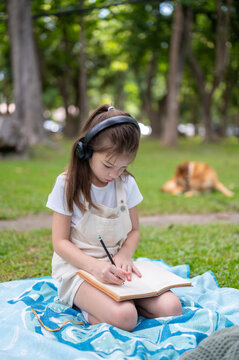 Young girl holding pencil writing in a notebook on picnic blanket while a dog play nearby in park.