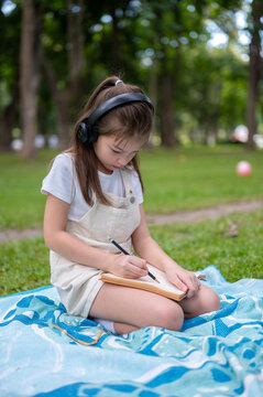 Young girl wearing headphones writing drawing in a notebook while sitting on picnic blanket in park