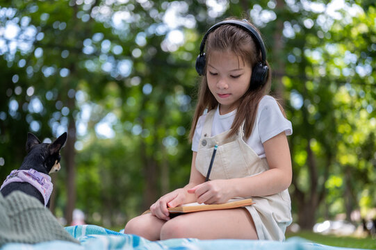 Young mixed race girl wearing headphones concentrates on writing drawing in notebook in the park.