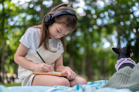 Young girl kid wearing headphones holding pencil writing in a notebook on blanket in the park.
