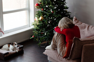 Naklejka premium Sad senior woman sitting alone on sofa hugging knees near christmas tree feeling lonely. Depression and solitude during holiday season concept. Mental health and grief at Christmas