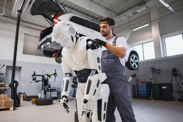 A man in overalls works on a humanoid robot in a car repair shop with a vehicle lifted overhead