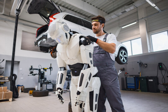 A man in overalls works on a humanoid robot in a car repair shop with a vehicle lifted overhead