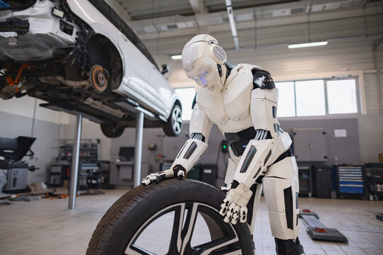 A humanoid robot with glowing blue eyes is carefully handling a car tire in a repair shop setting