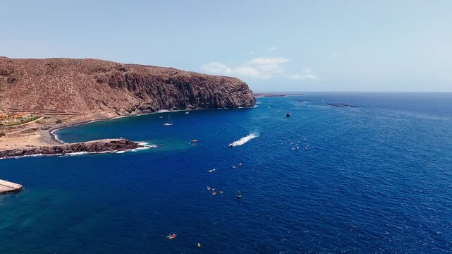 Wide drone view of the volcanic coast near Los Cristianos, with barren cliffs, rocky shoreline and clear blue ocean waters in Tenerife
