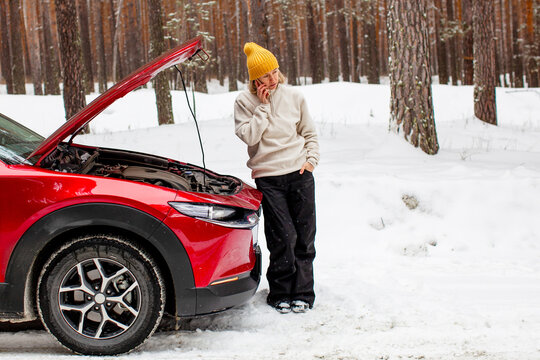 Woman standing near broken red car with open hood in snowy forest calling for roadside assistance on smartphone. Vehicle breakdown and emergency help problem in winter travel concept
