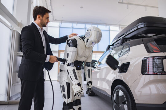 A man in a suit connects a charging cable to a humanoid robot next to an electric vehicle in a showroom