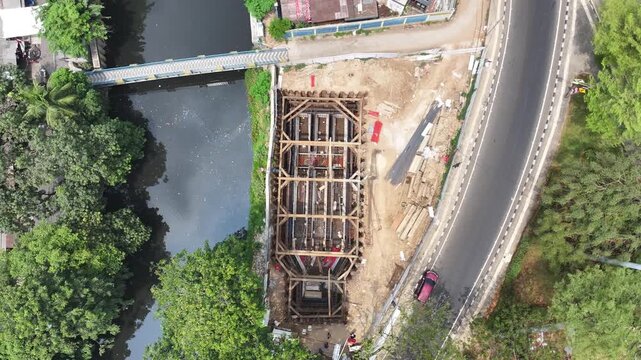 Workers build foundations near a calm river. Steel beams and wooden frames stand tall. Red pipes lie scattered on dirt ground. People move carefully through the construction site street construction