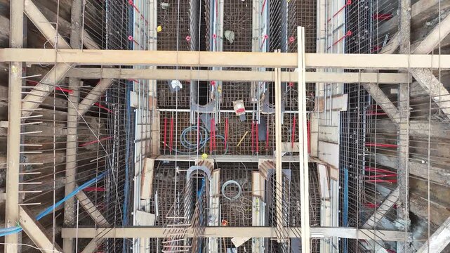 Workers build foundations near a calm river. Steel beams and wooden frames stand tall. Red pipes lie scattered on dirt ground. People move carefully through the construction site street construction