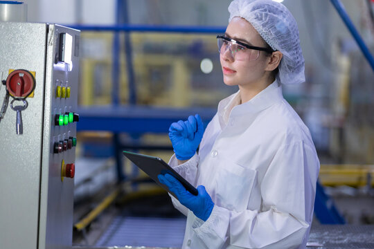 female worker wearing white safety uniform holding digital tablet checking machine control panel in manufacturing factory ensuring quality production
