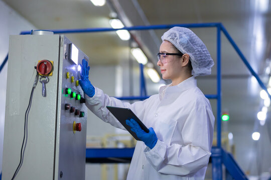 Caucasian female factory worker in white coat hairnet and blue glove operating industrial control panel holding digital tablet checking manufacturing process