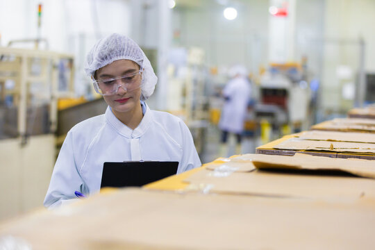 Asian woman factory worker performing quality inspection on manufacturing line wearing clean room uniform holding digital tablet checking cardboard box production process