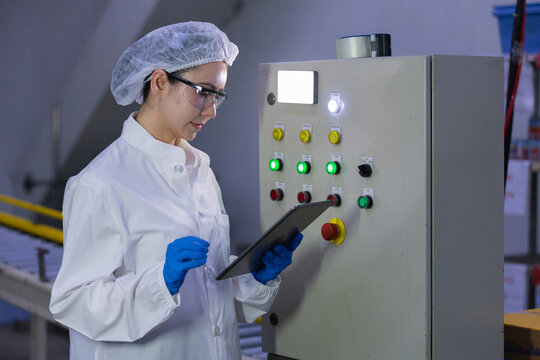 Female factory worker in white uniform holding digital tablet checking manufacturing control panel with focused expression ensuring quality standard during industrial production facility process