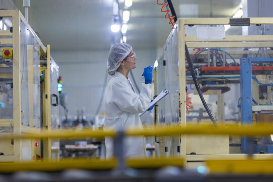 female factory worker wearing uniform and hairnet holding clipboard during inspection of clean room machinery showing attentive emotion while managing manufacturing plant production line