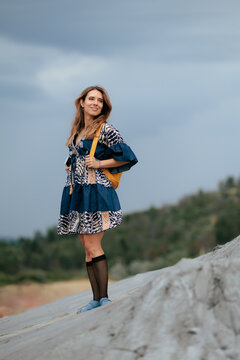 Smiling Young Woman Standing On Sand Dune Looking Up. Cheerful traveler wearing cool indian dress holds backpack straps stands outside thoughtfully 