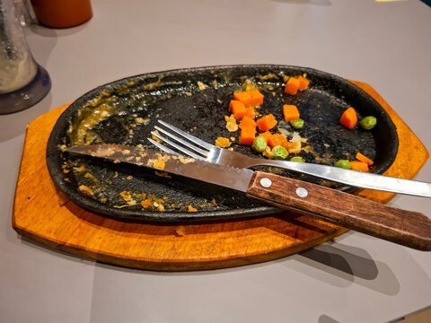 An empty sizzling plate with leftover vegetable bits, a fork, and a knife after a finished meal at a restaurant.