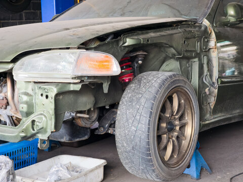 A green car raised on a jack stand in a garage with the front fender removed to work on red coilover suspension and wheel assembly.