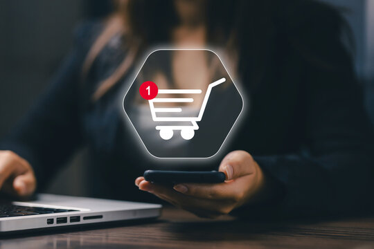 Businesswoman uses smartphone beside laptop with virtual shopping cart icon, representing ecommerce, online shopping, mobile payment, digital retail, customer order, and technology.
