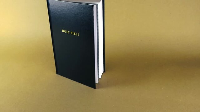 Holy Bible standing on a light brown background with golden text on the black cover and white pages. A slow camera movement glides from left to right, showcasing the sacred religious book
