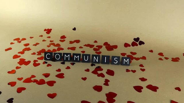 Conceptual representation showing the word communism written with black tiles on a light brown background, surrounded by scattered red shiny heart confetti in a smooth gliding shot