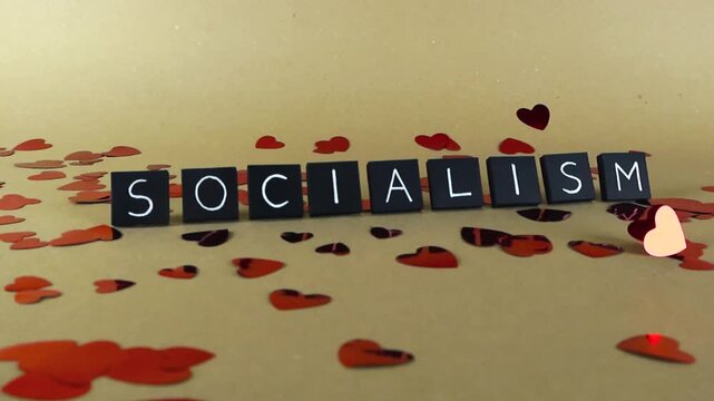 Black tiles spelling the word socialism surrounded by falling red heart confetti on a tan background, representing a political and economic concept with positive emotional connotations
