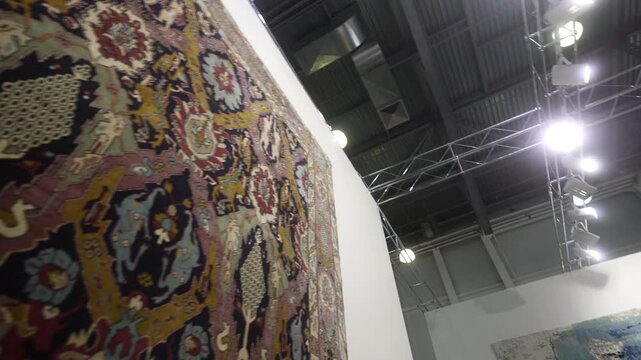 Low-angle pan of ornate hanging rug in gallery showing upper edge and industrial ceiling, with rug, carpet, textile, pattern detail, showing ornament, floral, geometric, woven context