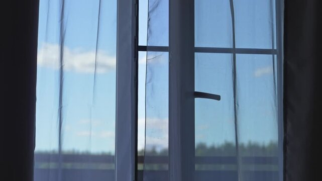 Gentle breeze blowing through an open window moves a delicate white tulle curtain, revealing a beautiful summer landscape with a blue sky, clouds, and a distant forest on a sunny day