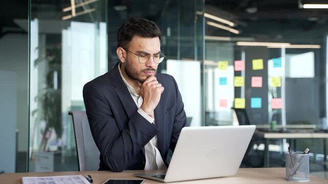 Serious thoughtful businessman in a formal suit thinking about problem solving while working on laptop sitting at workplace in business office. Focused entrepreneur is busy with a project on computer