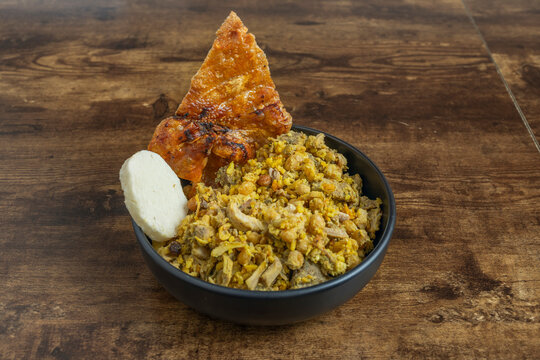 Black bowl of Colombian-style shredded meat and rice topped with crispy pork chicharr&oacute;n and a slice of white cheese.