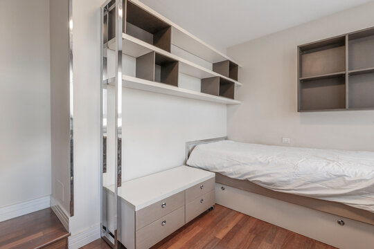 Compact bedroom with single bed, storage drawers, wall shelving cubbies, and mesh office chair by a sunlit window desk.