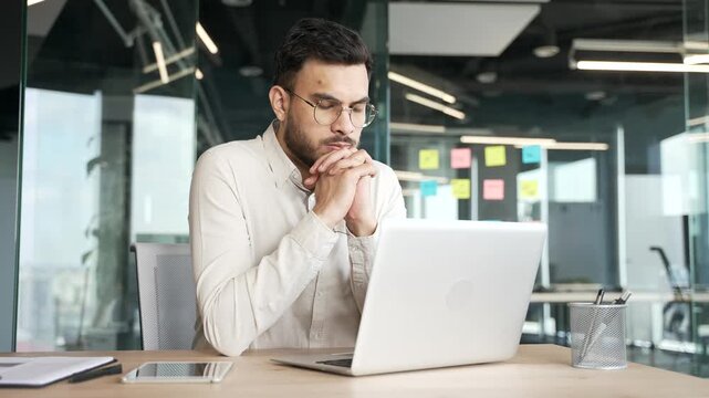 Thoughtful focused businessman thinking about problem solving while working on laptop sitting at desk at workplace in business office. Serious entrepreneur is busy with a startup project on computer